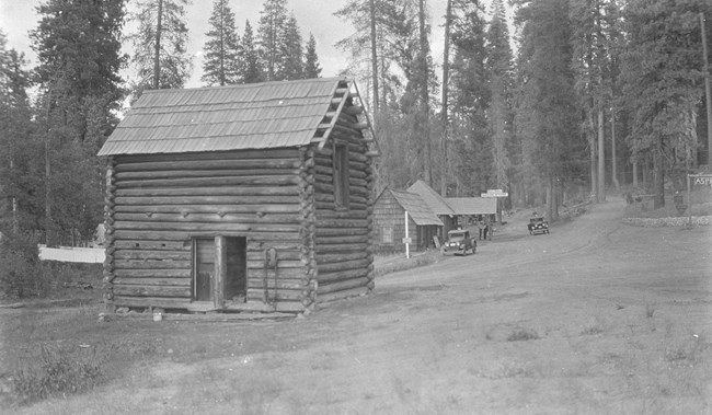 A wood log cabin stands next to a dirt road, another simple building and old cars are in the background