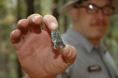 Ranger in background holding a piece of an obsidian spear point.