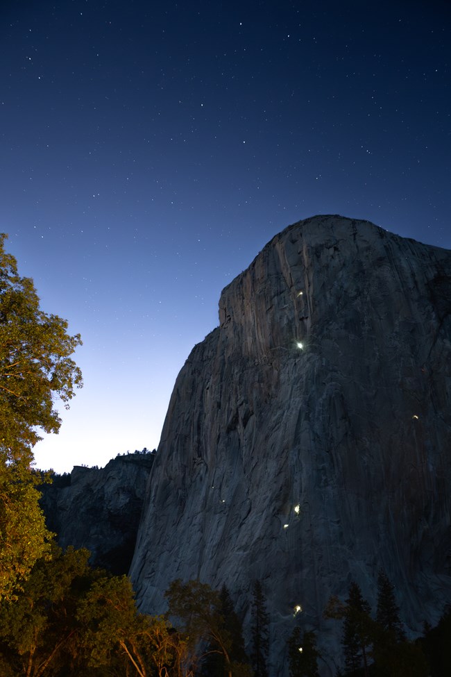 A dozen headlamps shine on a dramatic cliff face below a blue, starry sky.