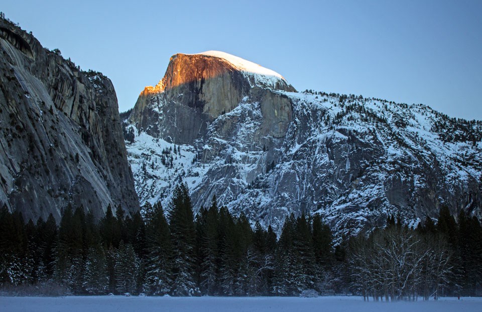 Orange light on the face of Half Dome with a snowy meadow in the foreground.