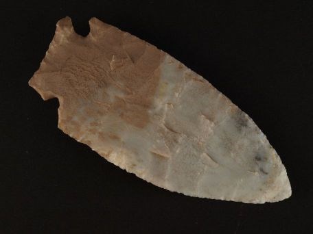 a stone knife blade made of chert in a large arrowhead shape, the rock is gray and reddish.