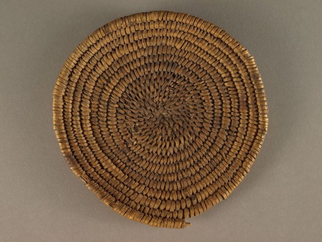 Flat woven basket in a spiral pattern with natural materials (dried yucca leaves around twigs).