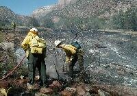 Two fire crew members work to put out a fire at the edge of a large burned area.