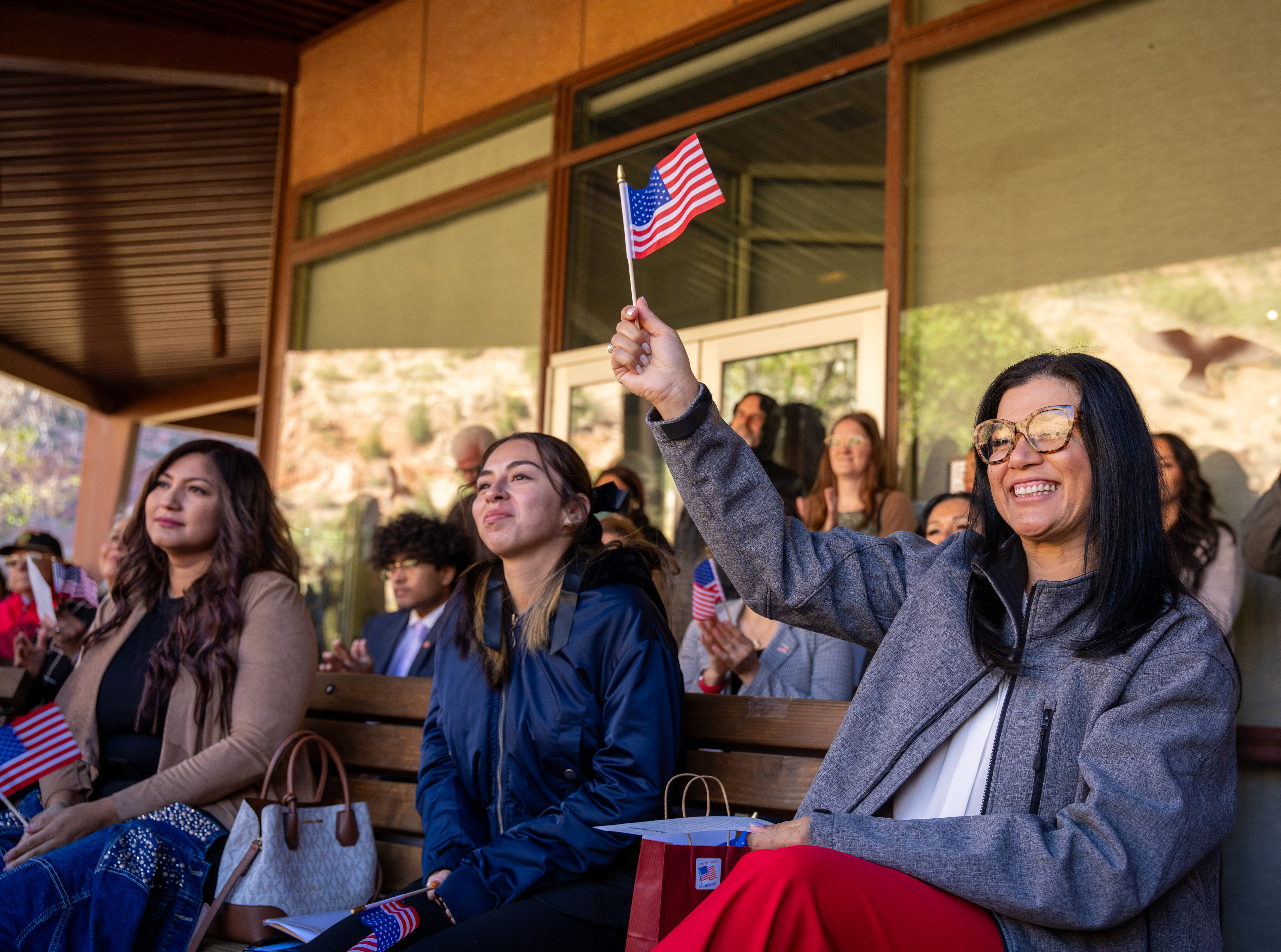 A woman sitting on a bench amongst a crowd smiling and waving an American Flag