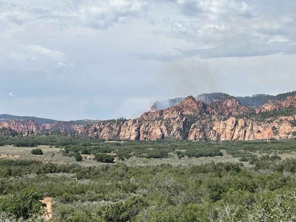 A green plain with red and orange sandstone cliffs in the background with smok coming from between the canyon