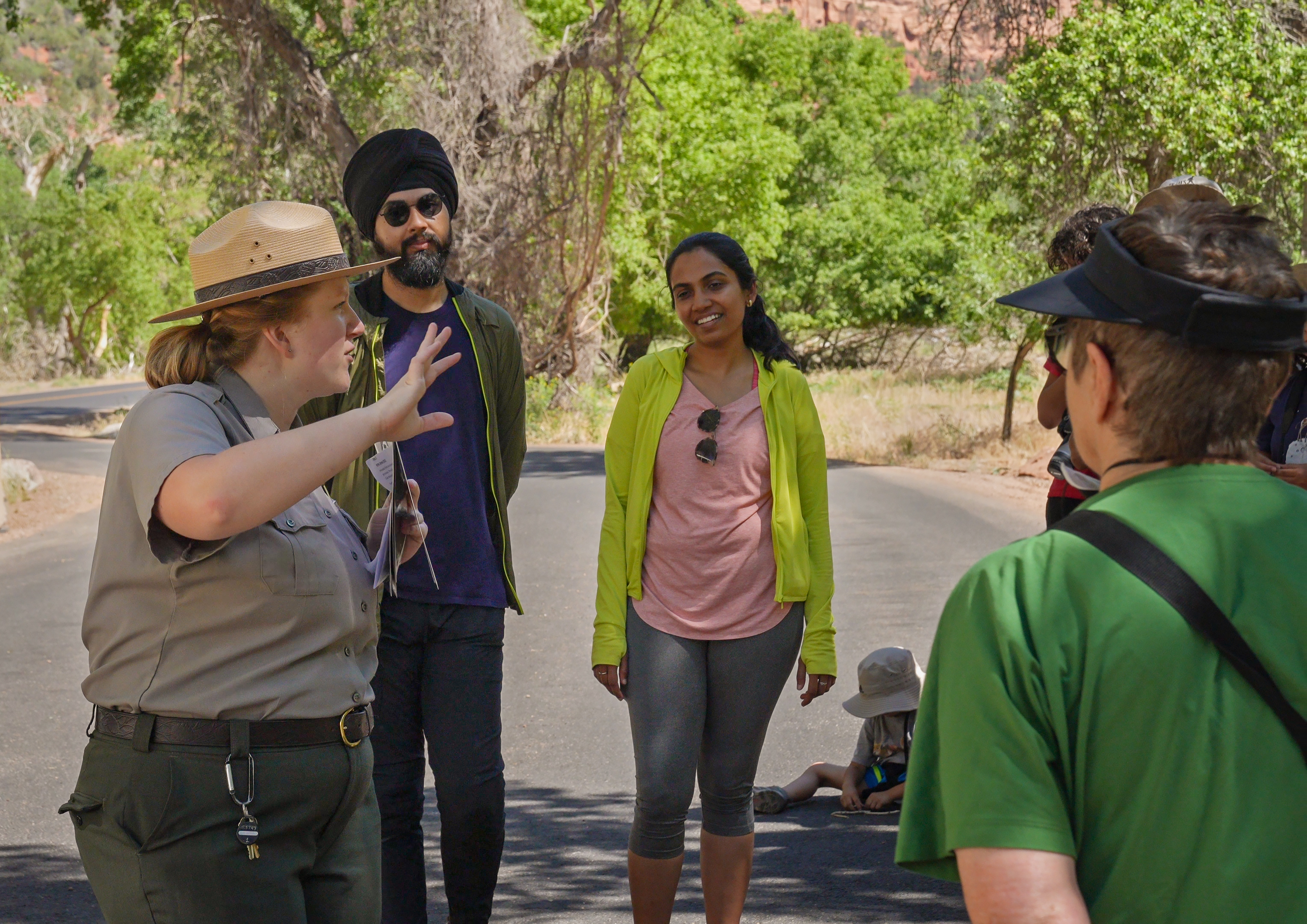 A park ranger talking to visitors at a road pullout with greenery and red sandstone in teh background