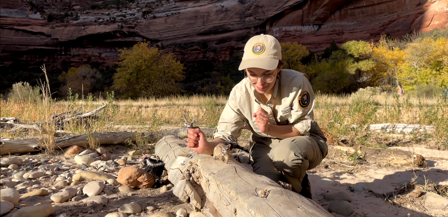 A young woman works with a stop-motion model of a deer. In the background, a river and tall cliffs are visible.