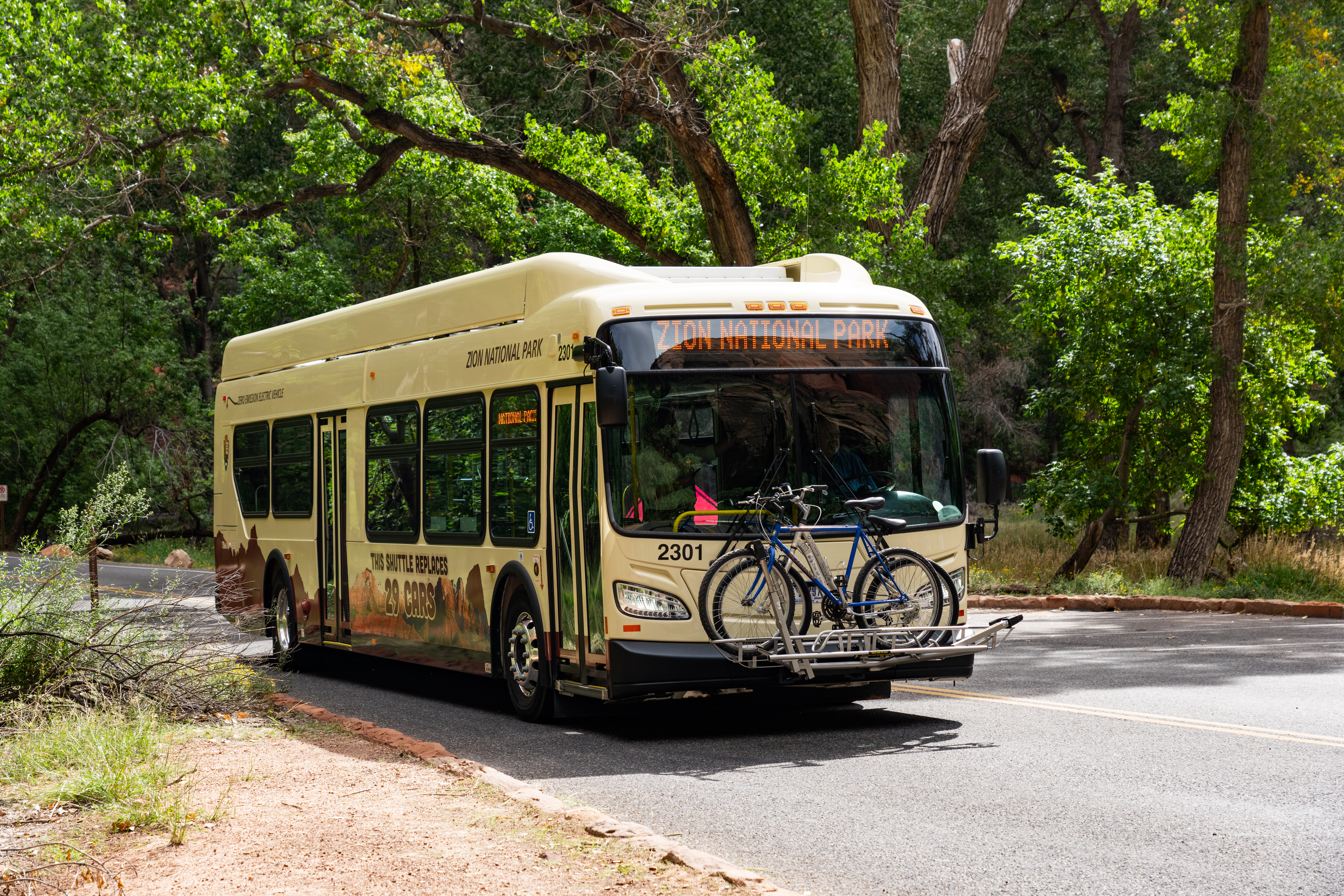 A light yellow electris bus with red graphics on the bottom and two bikes attached on the front with green trees in the background