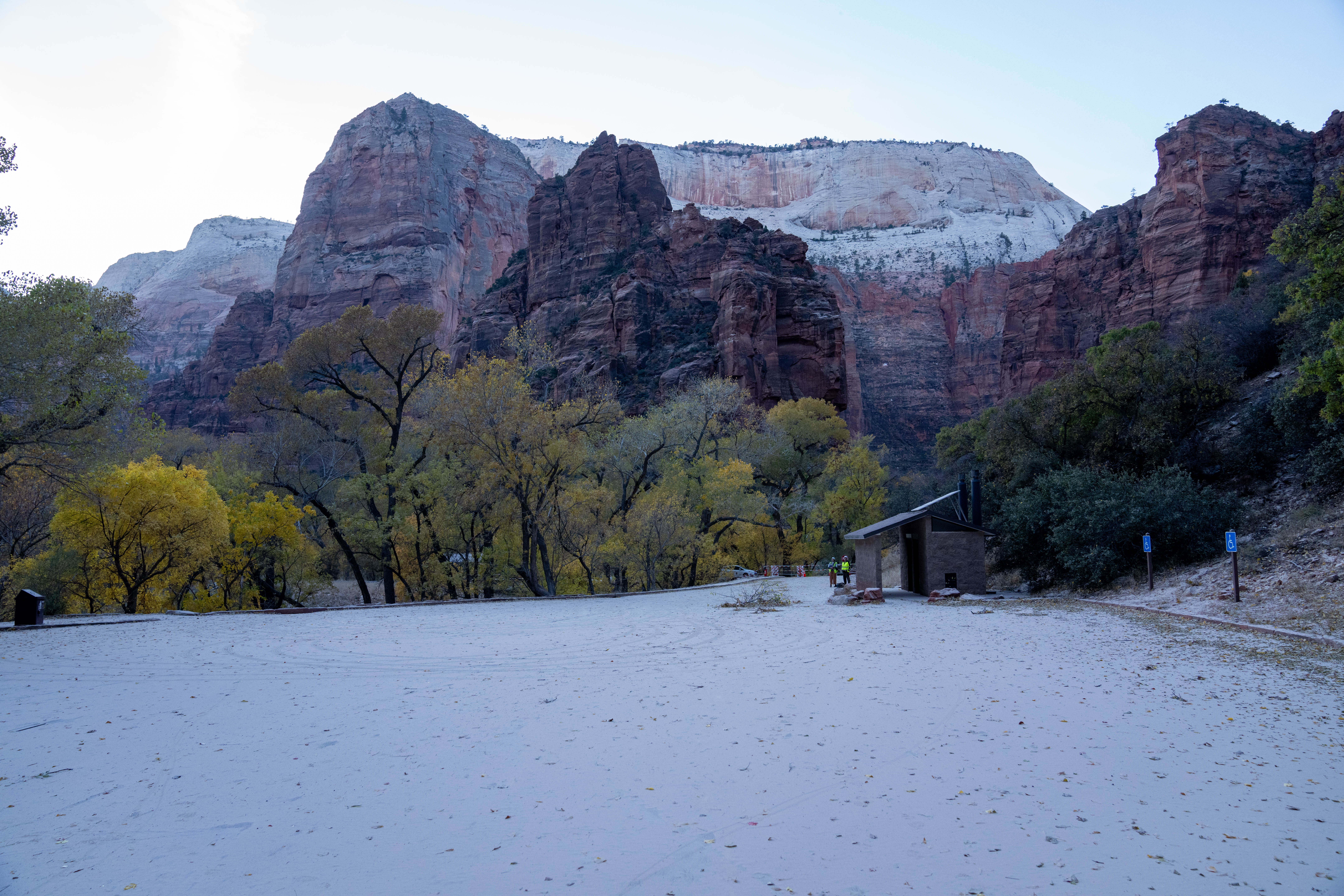 White dust covers a parking lot in front of a pit toilet building, trees, and towering red rock in the distance.