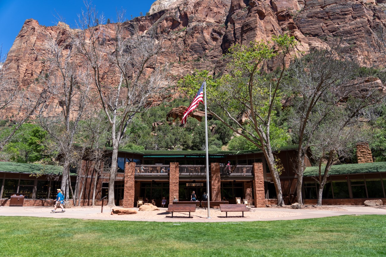Building with people standing outside, with a flag pole in teh foreground and sandstone cliffs and trees in teh background