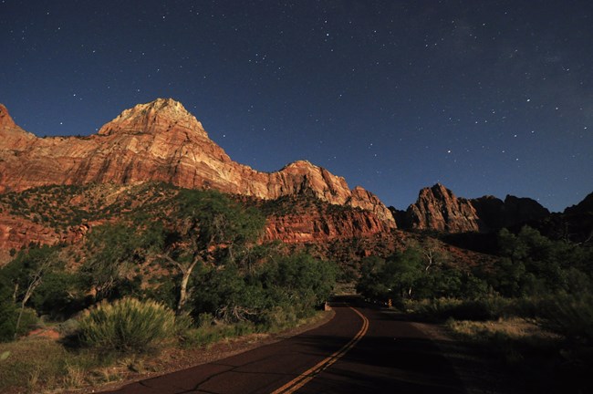 Red cliffs, a starry night sky, and an empty road stretching through the canyon.