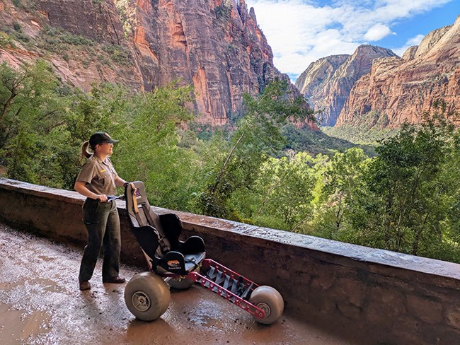 A park ranger holds the handlebars of an adaptive mobility wheelchair.